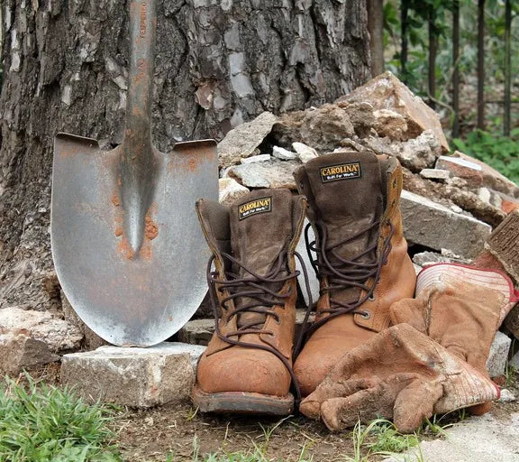 Brown work boots, gloves, and a shovel leaning against a tree with construction materials in the background.